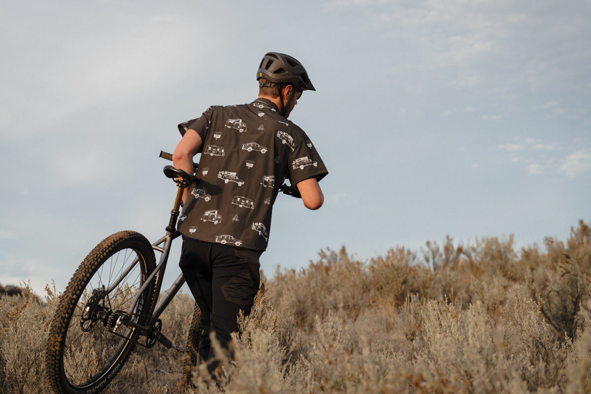 mountain biker carrying their chrome bike up a hill wearing black helmet and rowdee weekender shirt.