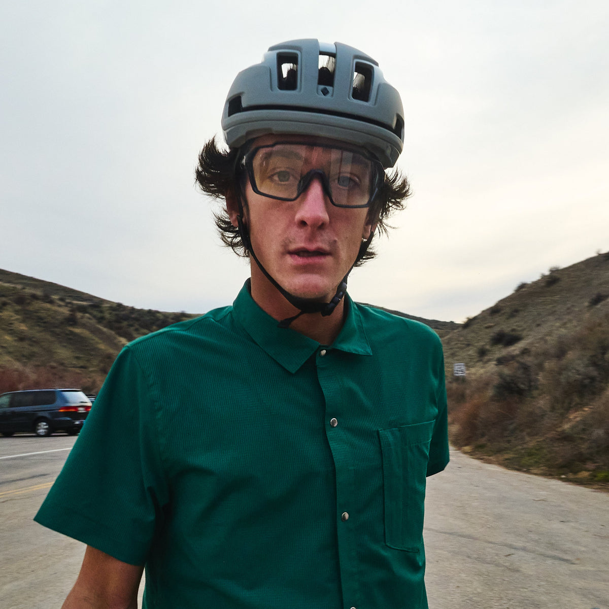 Person wearing a helmet and green shirt standing next to a bicycle on a road with mountains in the background