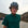Person wearing a helmet and green shirt standing next to a bicycle on a road with mountains in the background